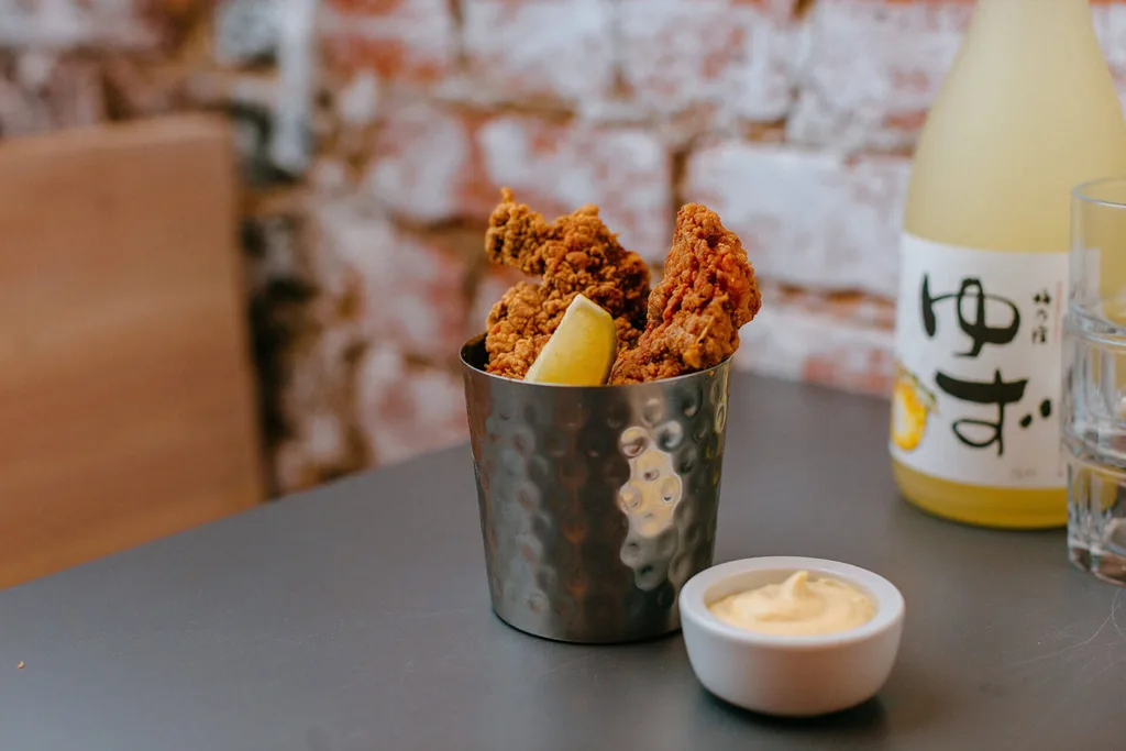 Kanada-Ya | Ramen Near Me | A metal cup filled with fried chicken and a slice of lemon sits on a gray table. Next to it is a small white bowl of dipping sauce. In the background, a bottle with Japanese writing is partially visible against a brick wall. | London