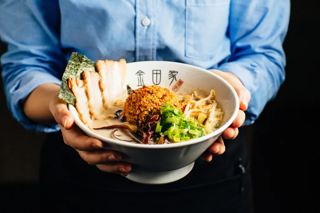 Kanada-Ya | Ramen Near Me | A person in a blue shirt holds a bowl of ramen with slices of meat, seaweed, greens, and a crispy topping, likely karaage. The ramen is in a white bowl with Asian characters, against a dark background. | London