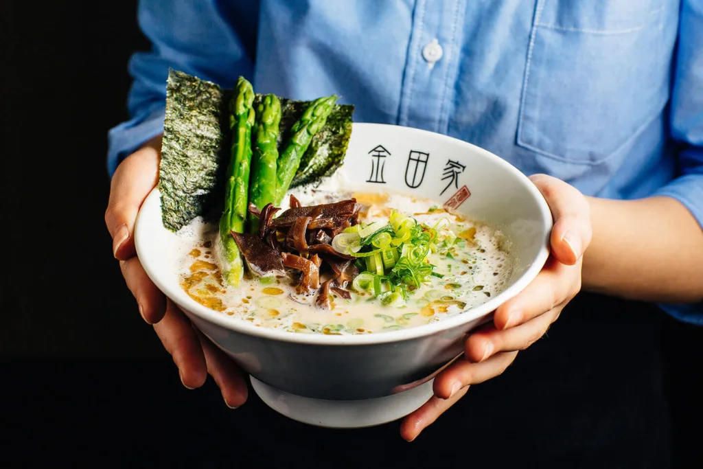 Kanada-Ya | Ramen Near Me | A person holds a bowl of ramen with creamy broth, topped with asparagus, seaweed, green onions, and sliced mushrooms. The person is wearing a blue shirt, and the bowl has Japanese characters on it. | London