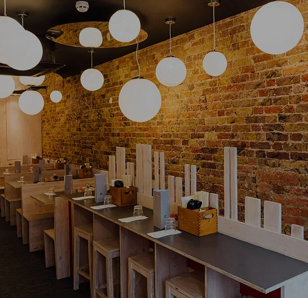 Kanada-Ya | Ramen Near Me | A cozy, modern restaurant interior with wooden tables and stools. Brick walls are adorned with spherical pendant lights, and tables have small decorative dividers and condiment holders. Warm lighting creates an inviting atmosphere. | London