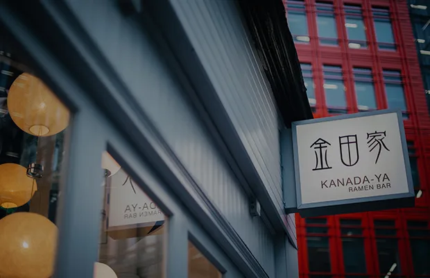 Kanada-Ya | Ramen Near Me | A close-up of the exterior of Kanada-Ya Ramen Bar, showing a sign with Japanese characters and the bars name in English. The building has large windows, with warm lights visible inside. A red building facade is visible in the background. | London