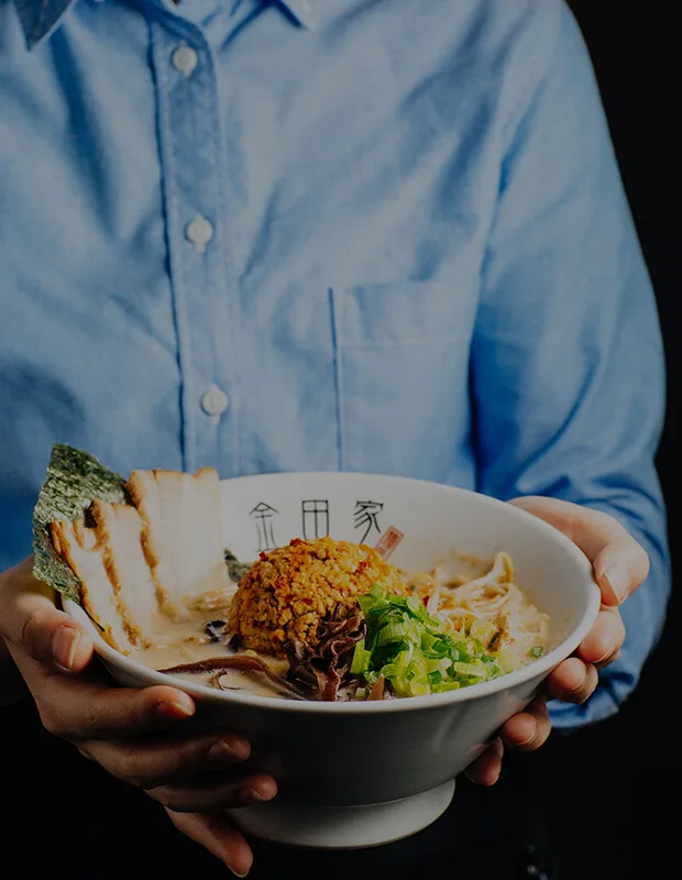 Kanada-Ya | Ramen Near Me | A person in a blue shirt holds a bowl of ramen, featuring sliced meat, a breaded topping, seaweed, mushrooms, and green onions. The bowl has a white interior with Japanese characters. | London