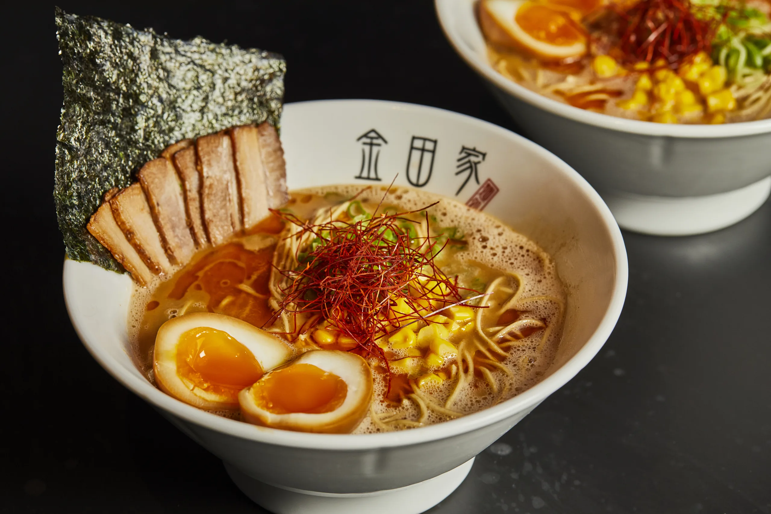 Kanada-Ya | Ramen Near Me | A bowl of ramen with sliced pork, seaweed, soft-boiled eggs, corn, and red pepper threads. The broth appears rich and is served in a white bowl with Japanese writing. Another bowl is partially visible in the background. | London